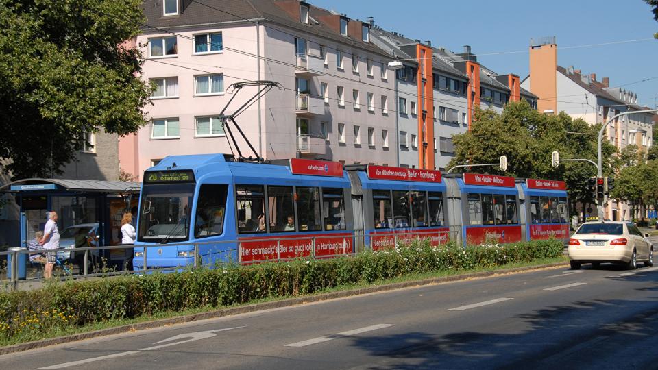 eine blaue Straßenbahn steht an einer Haltestelle, im Hintergrund sind mehrstöckige Wohnhäuser